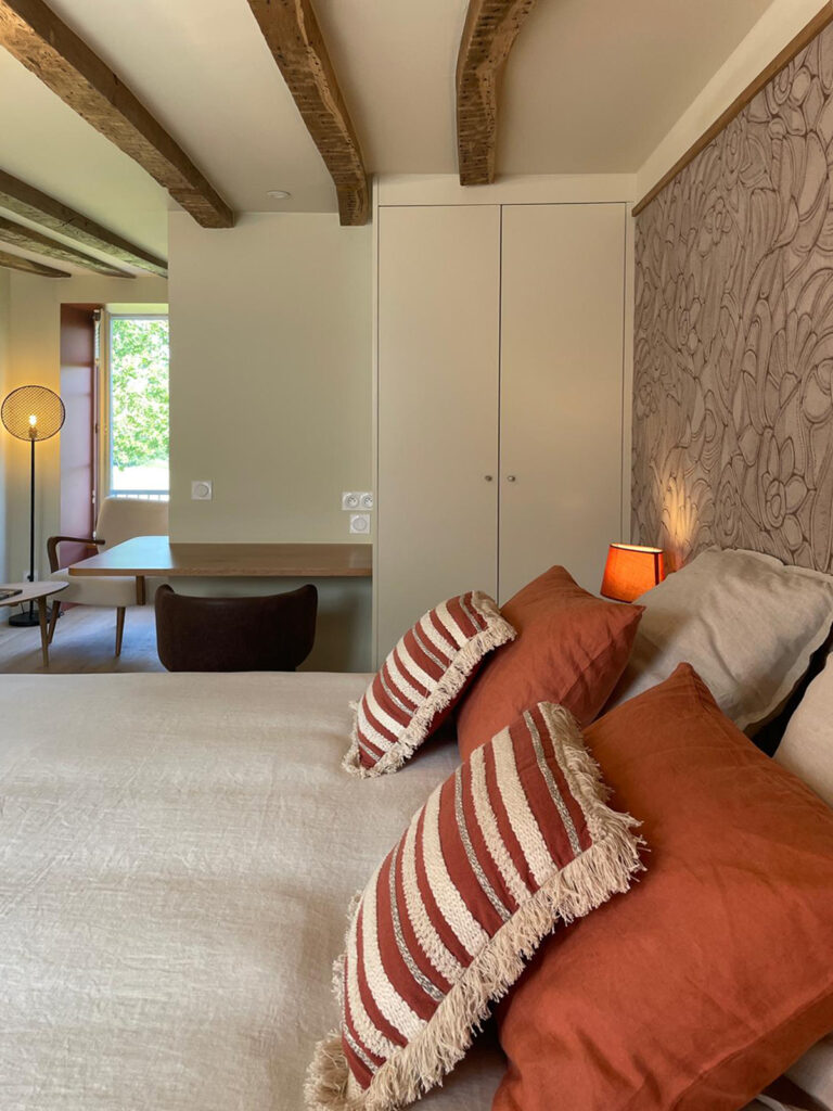 Chambre avec poutres en bois apparentes, lit avec coussins terracotta et bureau près de la fenêtre.