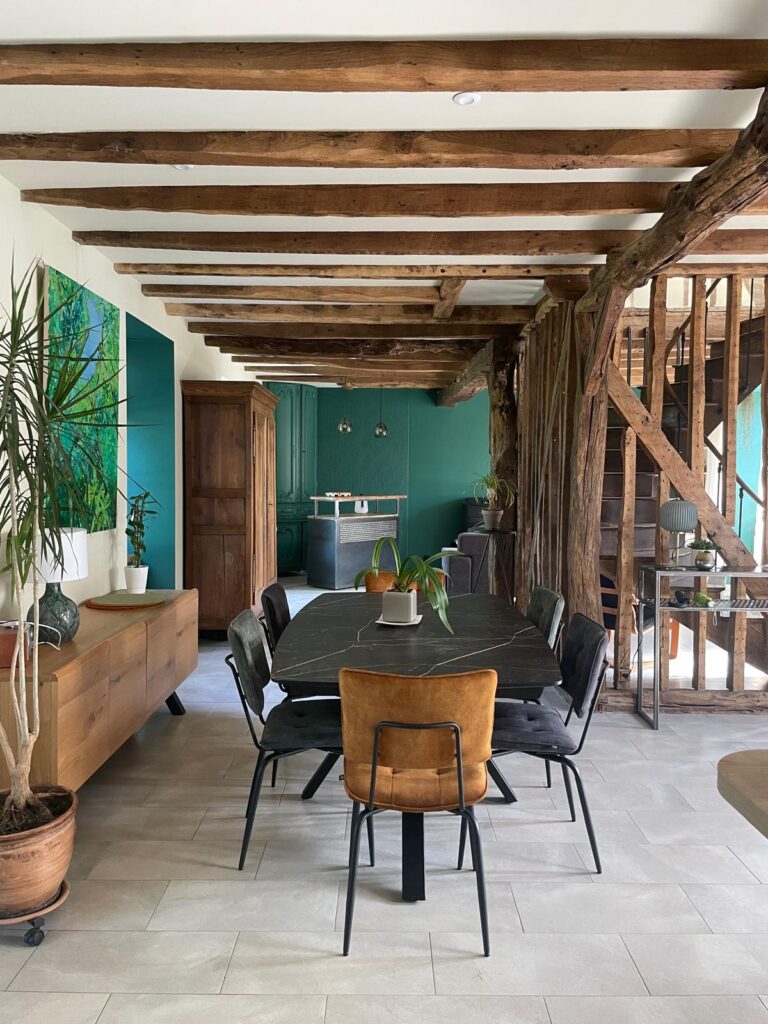 Salle à manger avec poutres en bois apparentes, table noire, chaises en velours, et murs peints en vert.